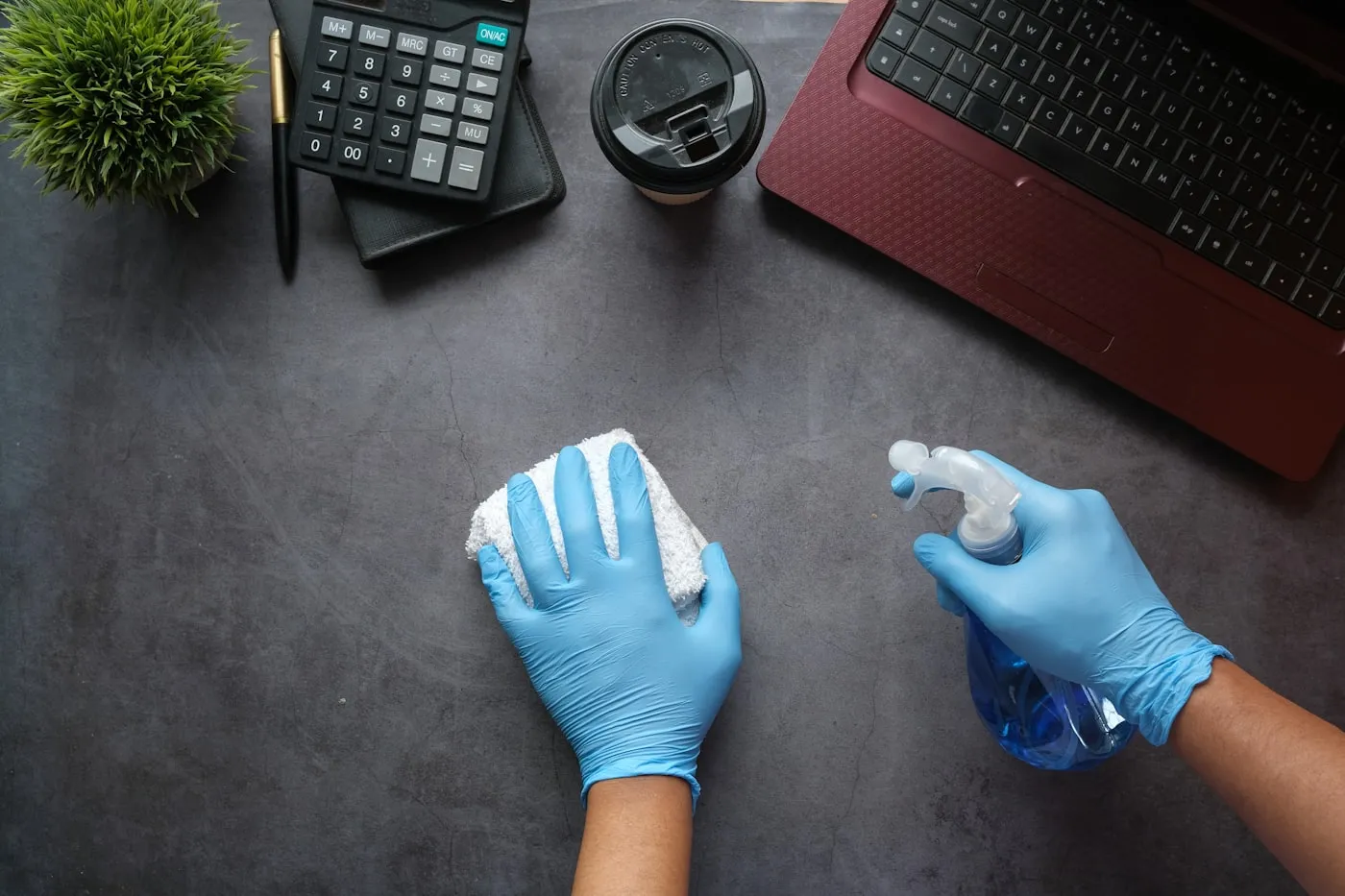 Gloved cleaner disinfecting an office desk beside a laptop